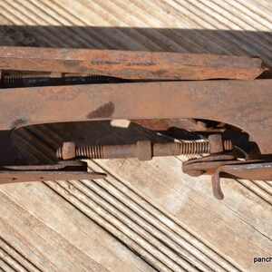 May include: A rusted metal ice skate blade assembly featuring a long, flat runner and a threaded adjustment rod with a central knurled nut. The iron components show significant oxidation and surface wear, with visible bolts and metal brackets.