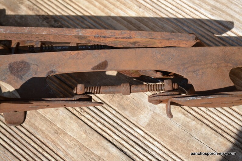 May include: A rusted metal ice skate blade assembly featuring a long, flat runner and a threaded adjustment rod with a central knurled nut. The iron components show significant oxidation and surface wear, with visible bolts and metal brackets.