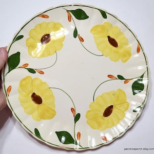 May include: A round, off-white ceramic plate with a scalloped edge. The plate is decorated with four large yellow sunflowers with brown centers and green leaves. The rim has a green border with orange accents.