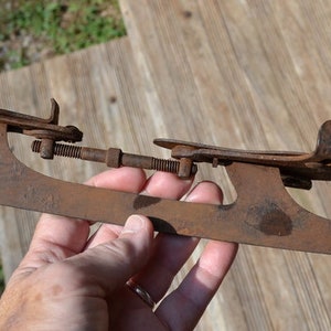 May include: A vintage, rusted metal ice skate blade featuring an adjustable screw mechanism and two metal foot plates. The skate has a weathered, brown oxidised finish and a long, flat blade design with curved ends, showing signs of age and wear.