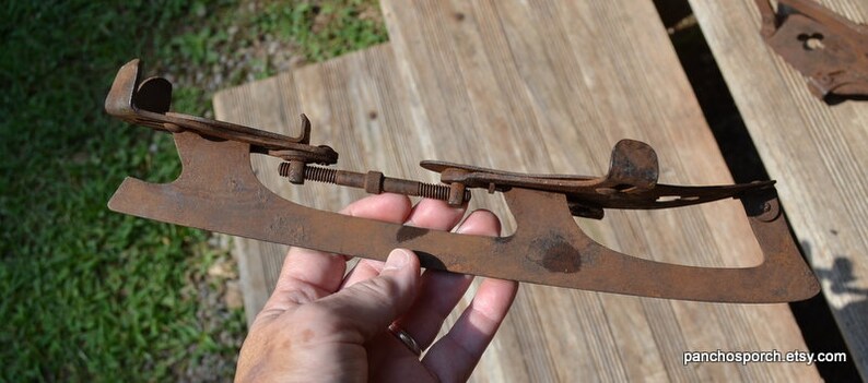May include: A vintage, rusted metal ice skate blade featuring an adjustable screw mechanism and two metal foot plates. The skate has a weathered, brown oxidised finish and a long, flat blade design with curved ends, showing signs of age and wear.