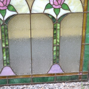 Op de afbeelding: Een glas-in-loodraam met een bloemenmotief. Het raam heeft drie panelen, elk met een roze roos. De panelen worden gescheiden door een dun, bruin frame. Het raam is geplaatst tegen een lichtbruine houten achtergrond.