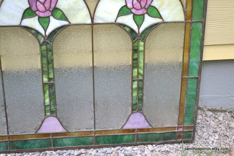 Op de afbeelding: Een glas-in-loodraam met een bloemenmotief. Het raam heeft drie panelen, elk met een roze roos. De panelen worden gescheiden door een dun, bruin frame. Het raam is geplaatst tegen een lichtbruine houten achtergrond.