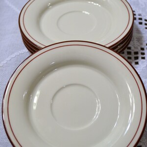 May include: A stack of off-white saucers with a thin, dark red border. The saucers have a slightly recessed center and are arranged on a white tablecloth. The image is well-lit, highlighting the smooth surface of the saucers.