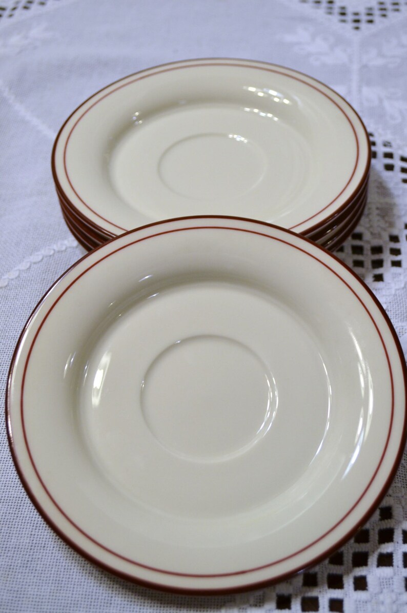 May include: A stack of off-white saucers with a thin, dark red border. The saucers have a slightly recessed center and are arranged on a white tablecloth. The image is well-lit, highlighting the smooth surface of the saucers.