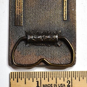 May include: Aged metal belt buckle with a bronze finish and decorative handle. The text reads: "This buckle is awarded to JWR employees with no lost time accidents in 1983. Make safety score in 84. Designed by David Sullivan."