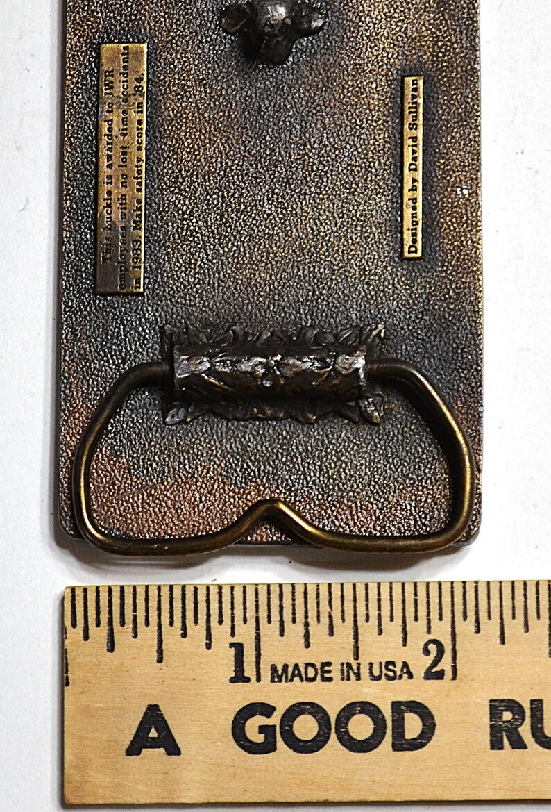 May include: Aged metal belt buckle with a bronze finish and decorative handle. The text reads: "This buckle is awarded to JWR employees with no lost time accidents in 1983. Make safety score in 84. Designed by David Sullivan."