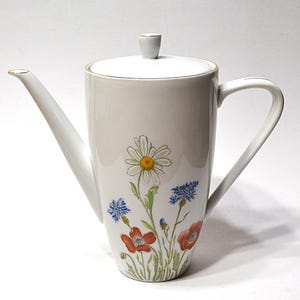 May include: White ceramic coffee pot with a long spout, handle, and lid. The pot is decorated with a floral design featuring daisies, poppies, and cornflowers. The rim and spout have a gold trim. A classic and elegant design.
