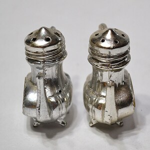 May include: Two vintage silver-colored salt and pepper shakers. Each shaker has a conical top with multiple holes, a ridged midsection, and a small handle. The shakers are on small feet and have a classic design.