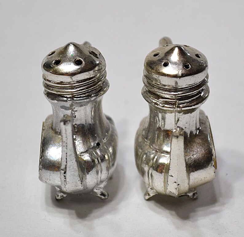 May include: Two vintage silver-colored salt and pepper shakers. Each shaker has a conical top with multiple holes, a ridged midsection, and a small handle. The shakers are on small feet and have a classic design.