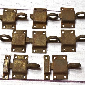 May include: A set of seven rectangular metal latches with a weathered, brownish-gold patina. Each latch features a central sliding bolt mechanism with a circular pull ring and four small mounting holes on the flat base plates for secure attachment.