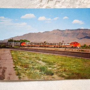 May include: A vintage postcard featuring a red and silver passenger train traveling along tracks through a desert landscape. In the background, large brown mountains rise under a bright blue sky with scattered white clouds. Green vegetation grows.