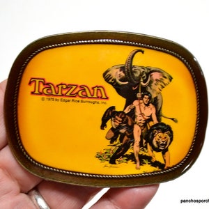 May include: An oval-shaped brass belt buckle featuring a yellow background with a graphic illustration of Tarzan, an elephant, and a lion. The text reads Tarzan and 1975 by Edgar Rice Burroughs, Inc. The buckle has a rope-style border design.