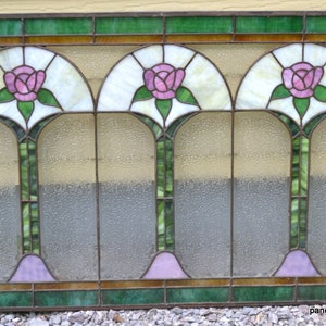Op de afbeelding: Een glas-in-loodraam met drie gebogen panelen met roze rozen en groene bladeren. Het raam heeft een lichtbruin houten frame en is geplaatst tegen een gele muur.