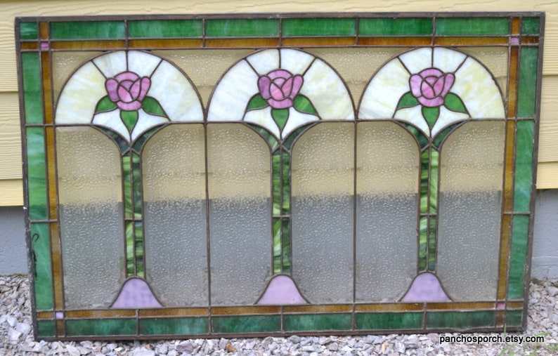 Op de afbeelding: Een glas-in-loodraam met drie gebogen panelen met roze rozen en groene bladeren. Het raam heeft een lichtbruin houten frame en is geplaatst tegen een gele muur.