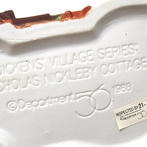 May include: A white ceramic base of a Dickens Village Series cottage figurine. The text "DICKENS VILLAGE SERIES" and "NICHOLAS NICKLEBY COTTAGE" are embossed on the base, along with the year 1988 and the Department 56 logo.