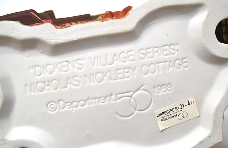 May include: A white ceramic base of a Dickens Village Series cottage figurine. The text "DICKENS VILLAGE SERIES" and "NICHOLAS NICKLEBY COTTAGE" are embossed on the base, along with the year 1988 and the Department 56 logo.