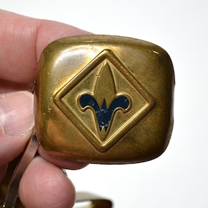 May include: A square-shaped brass neckerchief slide with rounded corners, featuring a raised diamond-shaped border surrounding a central dark blue fleur-de-lis emblem. The metal surface shows a weathered, aged patina with visible surface texture.