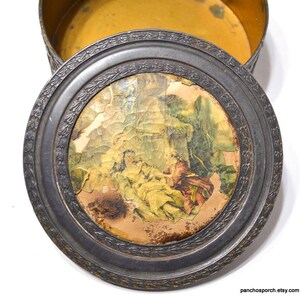 Op de afbeelding: Een antieke ronde metalen doos met een deksel met een vervaagde, gebarsten print van een klassieke sc&egrave;ne. Het deksel heeft een decoratieve, reli&euml;f rand. De binnenkant van de doos is gevoerd met gele stof.