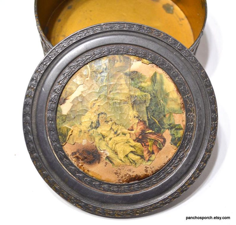 Op de afbeelding: Een antieke ronde metalen doos met een deksel met een vervaagde, gebarsten print van een klassieke sc&egrave;ne. Het deksel heeft een decoratieve, reli&euml;f rand. De binnenkant van de doos is gevoerd met gele stof.