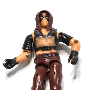 May include: A plastic action figure with a brown hood, black eye mask, and light skin tone. The figure wears a black chest plate, black shoulder armor, black gloves, and brown pants with a dark belt. The arms and legs are articulated at the joints.