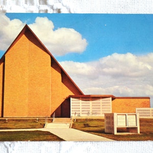 May include: A vintage postcard featuring a large, triangular-shaped brick building with a steep roofline. The structure has a light-coloured grid-patterned wall section, a concrete walkway leading to the entrance, and a small sign in the foreground.