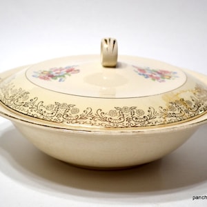 May include: A vintage cream-colored serving bowl with a lid. The bowl and lid are decorated with a gold floral pattern and have a floral design in the center.