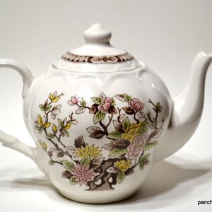 May include: White ceramic teapot with a floral design featuring pink, yellow, and green flowers on a brown branch. The teapot has a curved handle, a spout, and a lid with a decorative brown band. The Etsy shop name "panchosporch.etsy.com" is visible.
