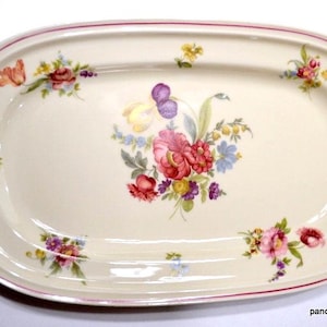 May include: An oval-shaped cream-colored ceramic serving platter featuring a central floral bouquet of pink, red, yellow, and purple flowers. The rim is decorated with a thin red line and smaller floral accents scattered around the edge.