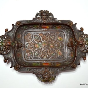 May include: An ornate, rectangular metal tray with a decorative, raised design. The tray has a dark brown finish with accents of green, red, and gold. The edges of the tray are intricately detailed with scrollwork and a central design featuring a grotesque face.