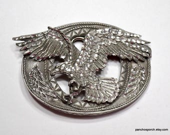 May include: Silver-toned belt buckle featuring a detailed eagle design with spread wings. The eagle is surrounded by a decorative border with a rope-like pattern.