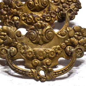 Peut inclure: Deux poignées de tiroir antiques en laiton avec des motifs floraux et des volutes ornés. Les poignées présentent une patine chaude et vieillie, avec des signes d'usure visibles, suggérant qu'elles sont vintage ou récupérées. Les poignées sont incurvées et présentent des détails complexes.