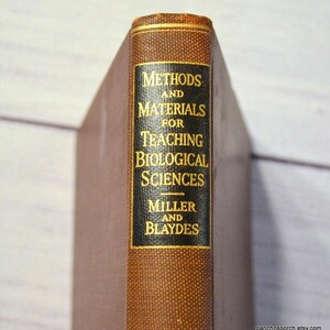 May include: A vintage book titled "Methods and Materials for Teaching Biological Sciences" by Miller and Blaydes. The spine is brown with gold lettering on a black background. The top edge of the book is gold.
