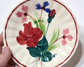 May include: A small, round, off-white ceramic plate with a scalloped edge and a red border. The plate is decorated with a hand-painted floral design featuring red, pink, and blue flowers, green leaves, and stems.