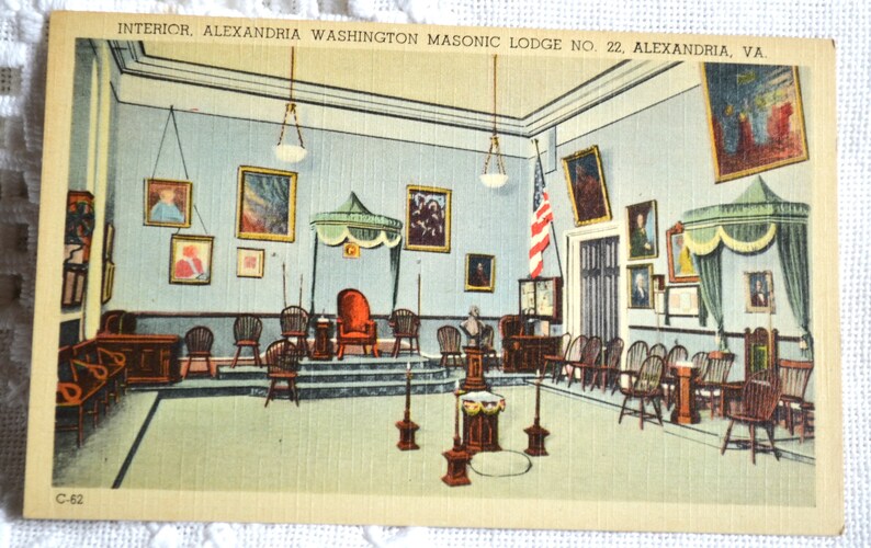 May include: A vintage postcard depicting the interior of the Alexandria Washington Masonic Lodge No. 22 in Alexandria, Virginia. The lodge room features a raised platform with a throne-like chair, surrounded by rows of chairs and a large American flag.