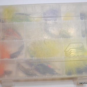 May include: A clear plastic Plano tackle box filled with colorful fishing flies. The flies are various colors, including orange, yellow, and black. The box is divided into compartments and has a Plano logo on the lid.