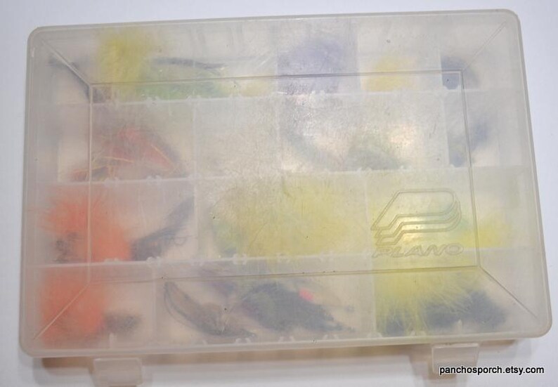 May include: A clear plastic Plano tackle box filled with colorful fishing flies. The flies are various colors, including orange, yellow, and black. The box is divided into compartments and has a Plano logo on the lid.