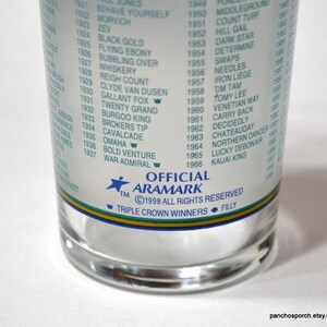 Pu&ograve; includere: Un bicchiere trasparente con un elenco di nomi e date stampati sul lato. Il bicchiere ha una fascia blu, verde e gialla vicino al fondo. Il testo include "OFFICIAL ARAMARK" e "TRIPLE CROWN WINNERS".