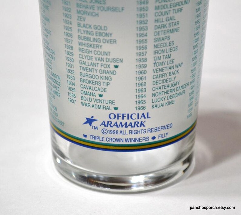 Pu&ograve; includere: Un bicchiere trasparente con un elenco di nomi e date stampati sul lato. Il bicchiere ha una fascia blu, verde e gialla vicino al fondo. Il testo include "OFFICIAL ARAMARK" e "TRIPLE CROWN WINNERS".