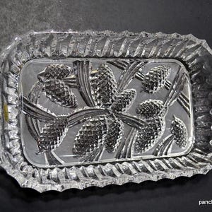 May include: A rectangular clear glass serving tray featuring a molded relief pattern of pinecones and pine needles on the base. The tray has integrated handles on both ends and a decorative serrated edge along the entire perimeter of the rim.