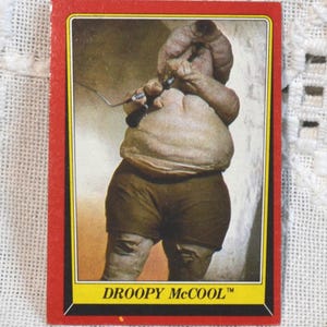 May include: A vintage trading card featuring Droopy McCool from Star Wars. The card has a red border with a yellow and black inner border. The image shows Droopy McCool in shorts, holding a musical instrument. The text "DROOPY McCOOL™" is at the bottom.