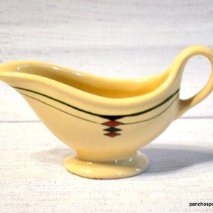 May include: Cream-colored gravy boat with a handle and a flared spout. The boat is decorated with black and green lines and a geometric pattern in red, black, and white. The gravy boat sits on a small, round base.