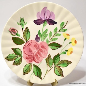 May include: Cream-colored ceramic plate with a ruffled edge, hand-painted with a floral design. The design features a large pink rose, purple flower, yellow flowers, and green leaves. The plate is supported by a gold-colored stand.