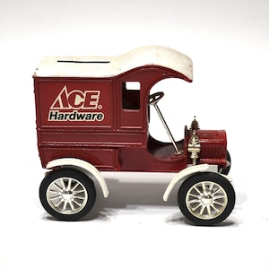 May include: A vintage-style red and white metal delivery truck coin bank featuring the Ace Hardware logo on the side. The vehicle has a white roof with a coin slot, silver-colored wheels with black tires, and a small steering wheel inside the cab.