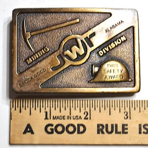 May include: A rectangular bronze-coloured belt buckle with the text "MINING BROOKWOOD" and "ALABAMA DIVISION". It features a pickaxe, the letters "JW", a hard hat, and the text "1988 SAFETY AWARD".