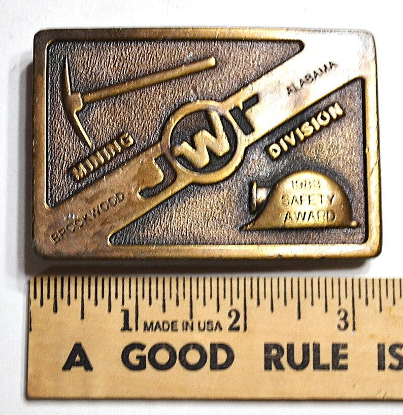 May include: A rectangular bronze-coloured belt buckle with the text "MINING BROOKWOOD" and "ALABAMA DIVISION". It features a pickaxe, the letters "JW", a hard hat, and the text "1988 SAFETY AWARD".