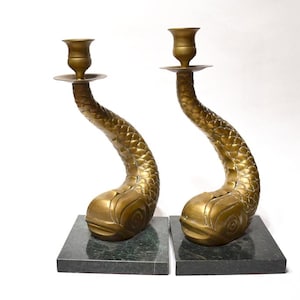 May include: Two antique brass candle holders with a fish tail design, set on dark green marble bases. The gold-coloured candle holders have a textured surface. The bases are square.