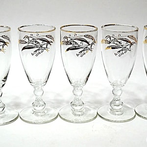 May include: Five clear champagne glasses with gold rims and a black and gold wheat design. Each glass has a round base and a decorative stem. The glasses are arranged in a row.