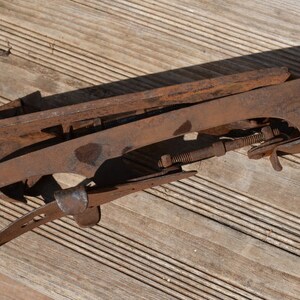 May include: A pair of vintage metal ice skates featuring a rusted brown finish, long blades, and adjustable threaded rods with nuts for tightening. The skates have a distinct clover-shaped cutout on the side frame and a curved heel support.