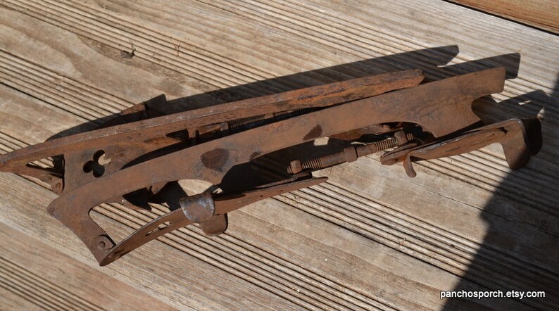 May include: A pair of vintage metal ice skates featuring a rusted brown finish, long blades, and adjustable threaded rods with nuts for tightening. The skates have a distinct clover-shaped cutout on the side frame and a curved heel support.